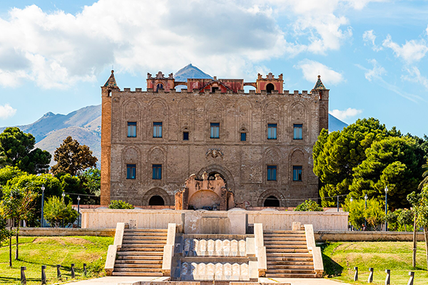 Storico palazzo in pietra con scalinata monumentale, fontana e giardini, sullo sfondo montagne e cielo nuvoloso.